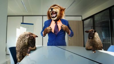 A lion dressed in a business suit roars as two sheep sit awkwardly at the boardroom table.