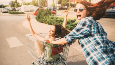 A laughing woman pushes her friend, who has her arms outstretched, in a supermarket trolley.