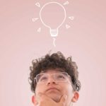 A youthful man looks up thoughtfully at a light bulb above his head.