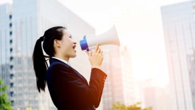 A woman standing among high rises shouts news through a megaphone.