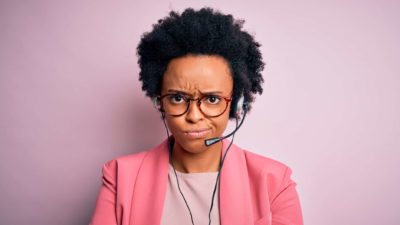 A well-dressed woman wearing a headset and mic looks grumpily at the camera.