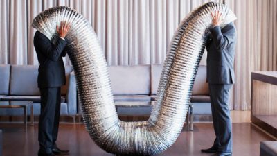 Two men in suits stand with their heads in either side of a big drooping silver ducting tube, trying to communicate.