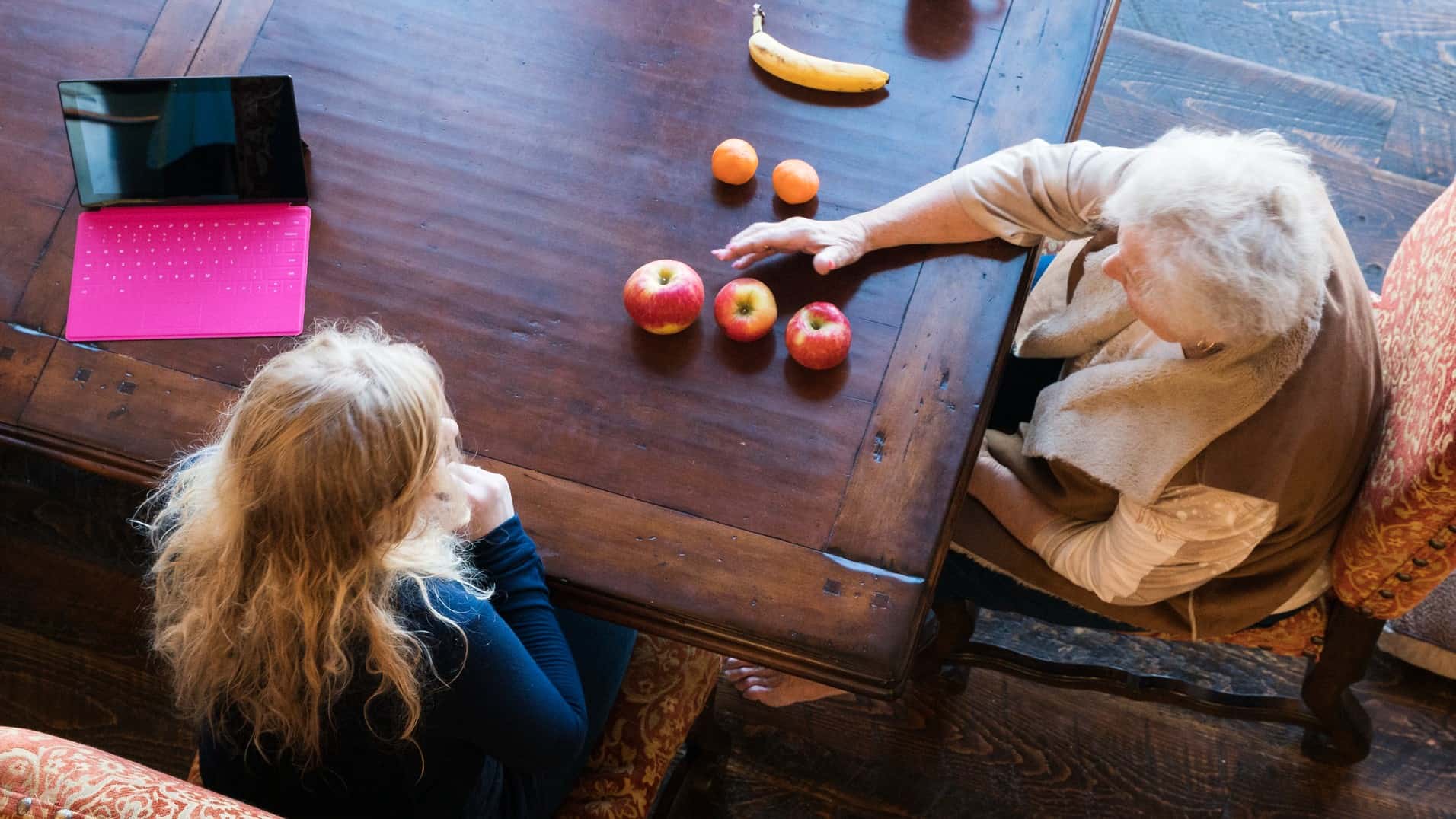 Looking down on an elderly woman teaching a younger woman using fruit, comparing apples with apples.
