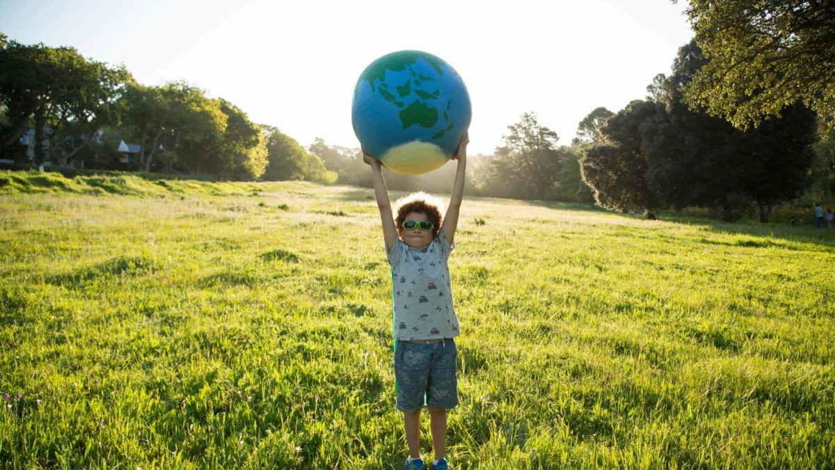 A young boy standing in a grassy field surrounded by trees holding a world globe over his head.