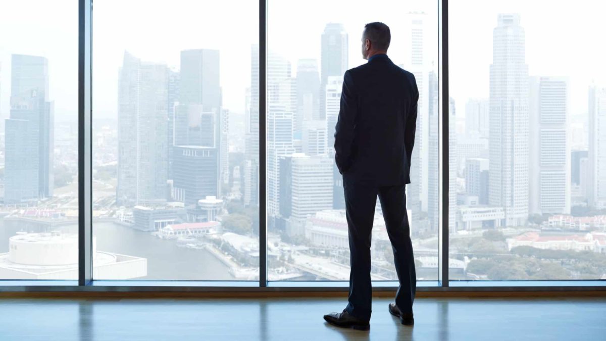 An executive stands looking out a glass window over the city.