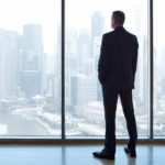 An executive stands looking out a glass window over the city.