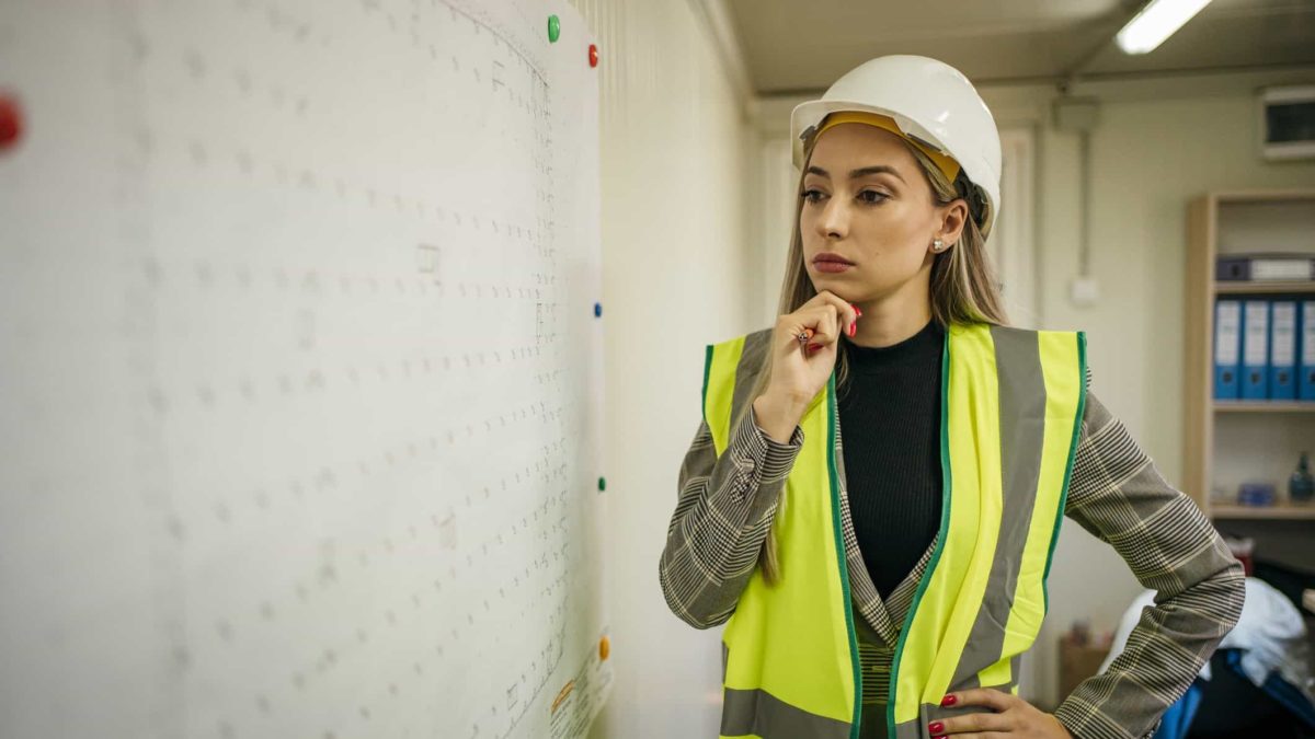 A female construction project manager in a hi vis vest and hard hat considers progress on a chart on the wall.