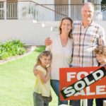 a family stands together behind a sold sign with their new house in the background.