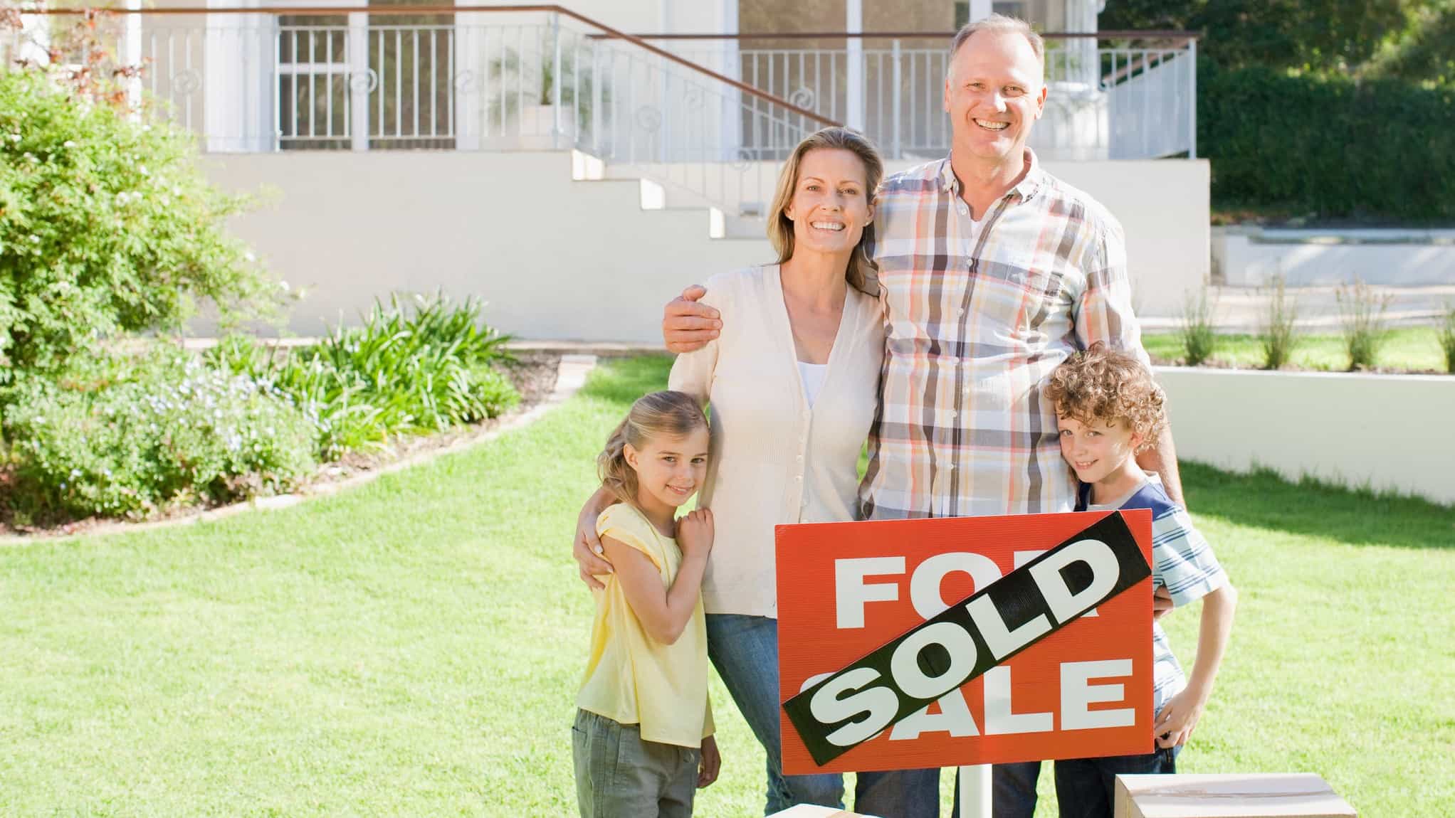 a family stands together behind a sold sign with their new house in the background.