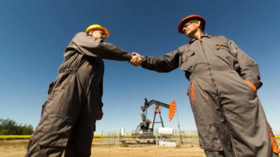 Two Santos oil workers with hard hats shake hands in the foreground of oil equipment.