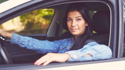 a woman sits in the driver's seat of a car with her arm resting on the door with a small smile on her face, looking out of the car.