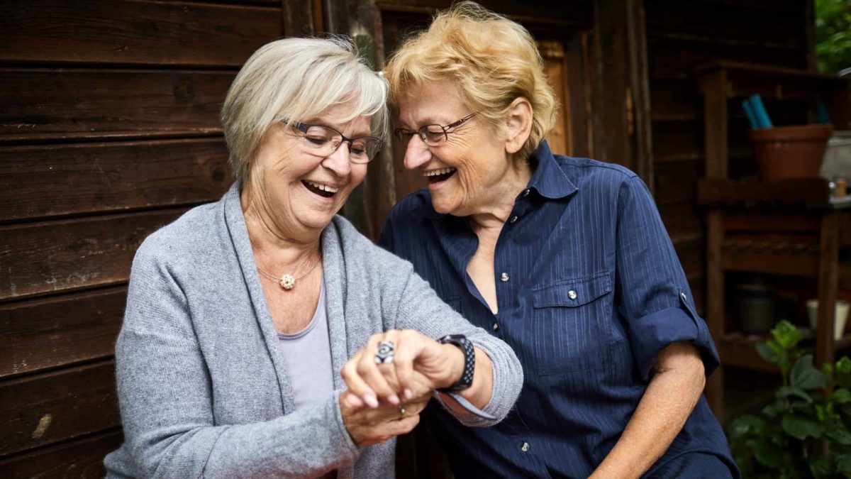 two women laugh as one talks into a watch style wearable communication device on her wrist.