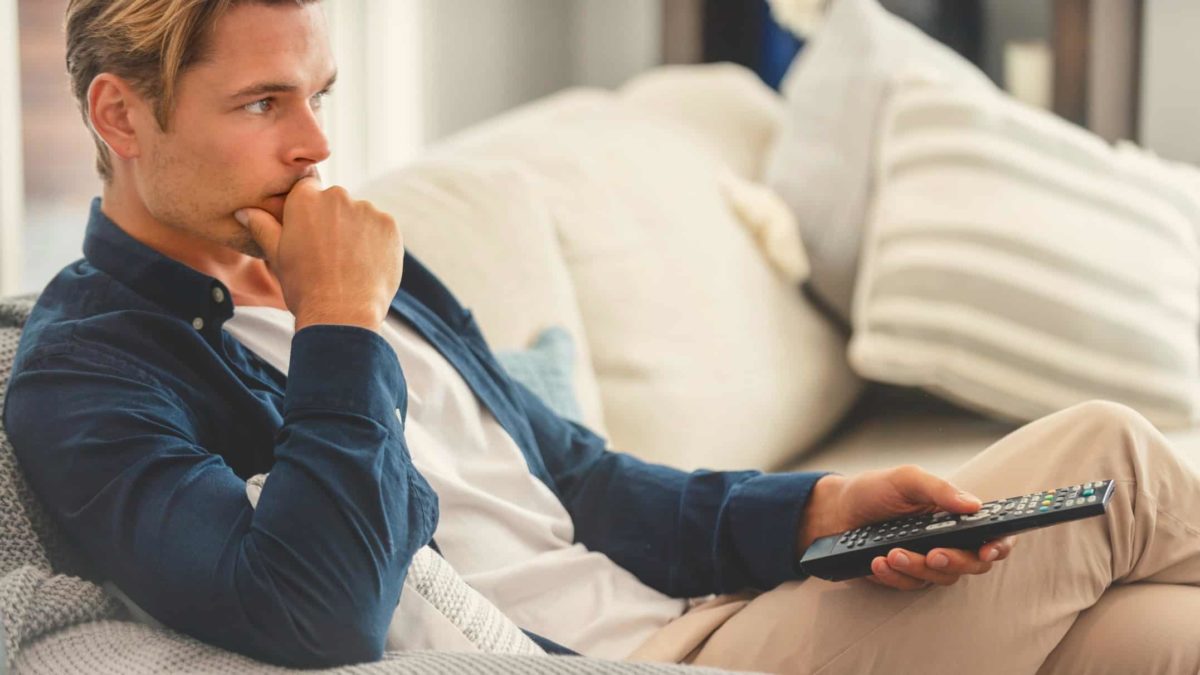 A man looks sad and reflective as he sits on his sofa with television remote control in hand.