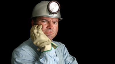 an unhappy miner poses with gloved hand on face wearing a hard hat with a light and frowning.