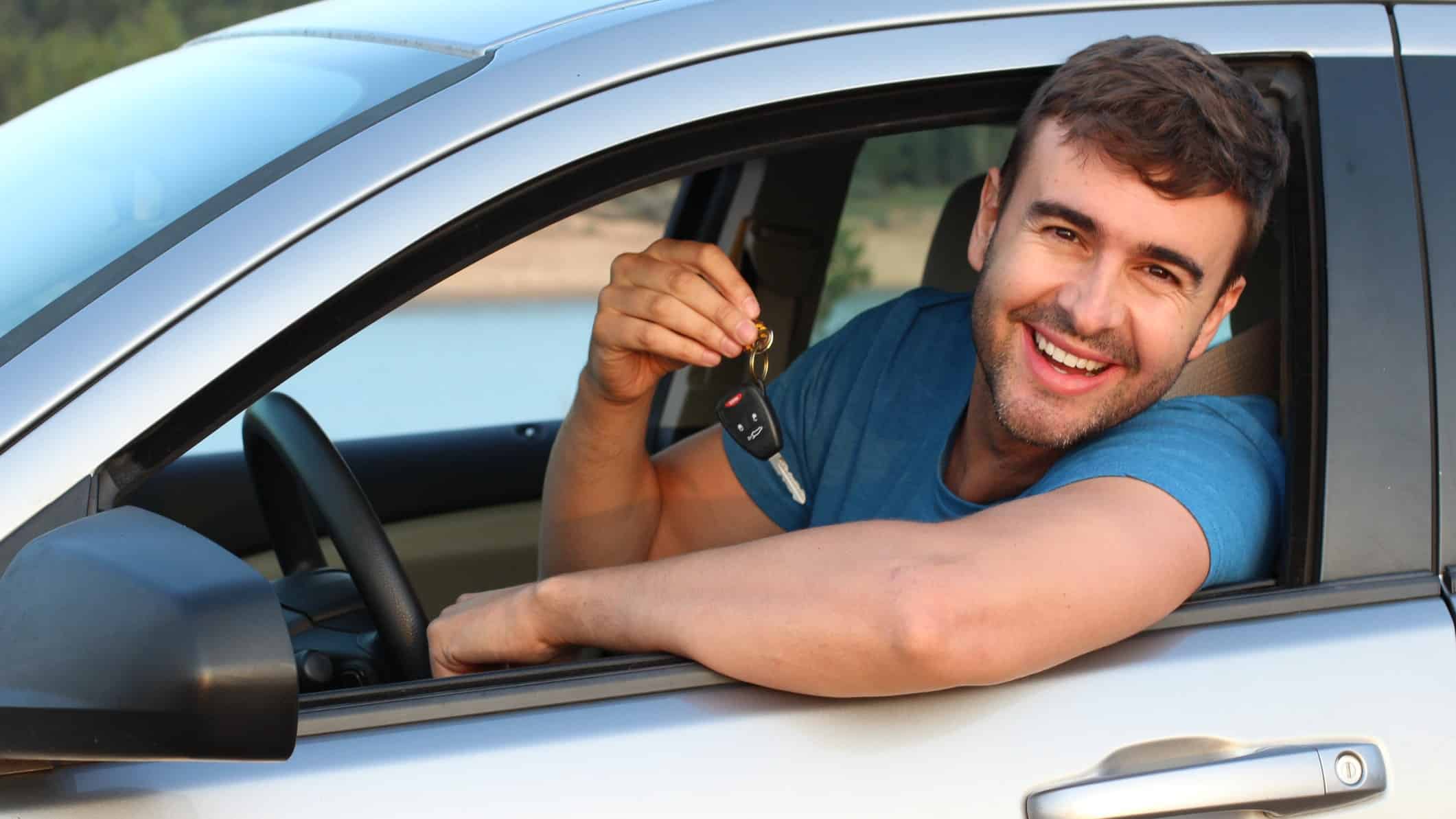 a smiling man leans out his car window, car keys in hand.