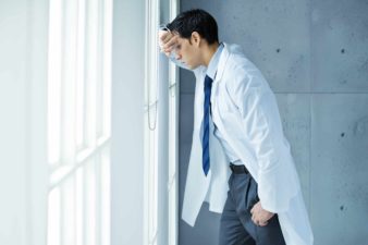 a doctor in white coat slumps against a window, head on hand, gazing down in dejection.