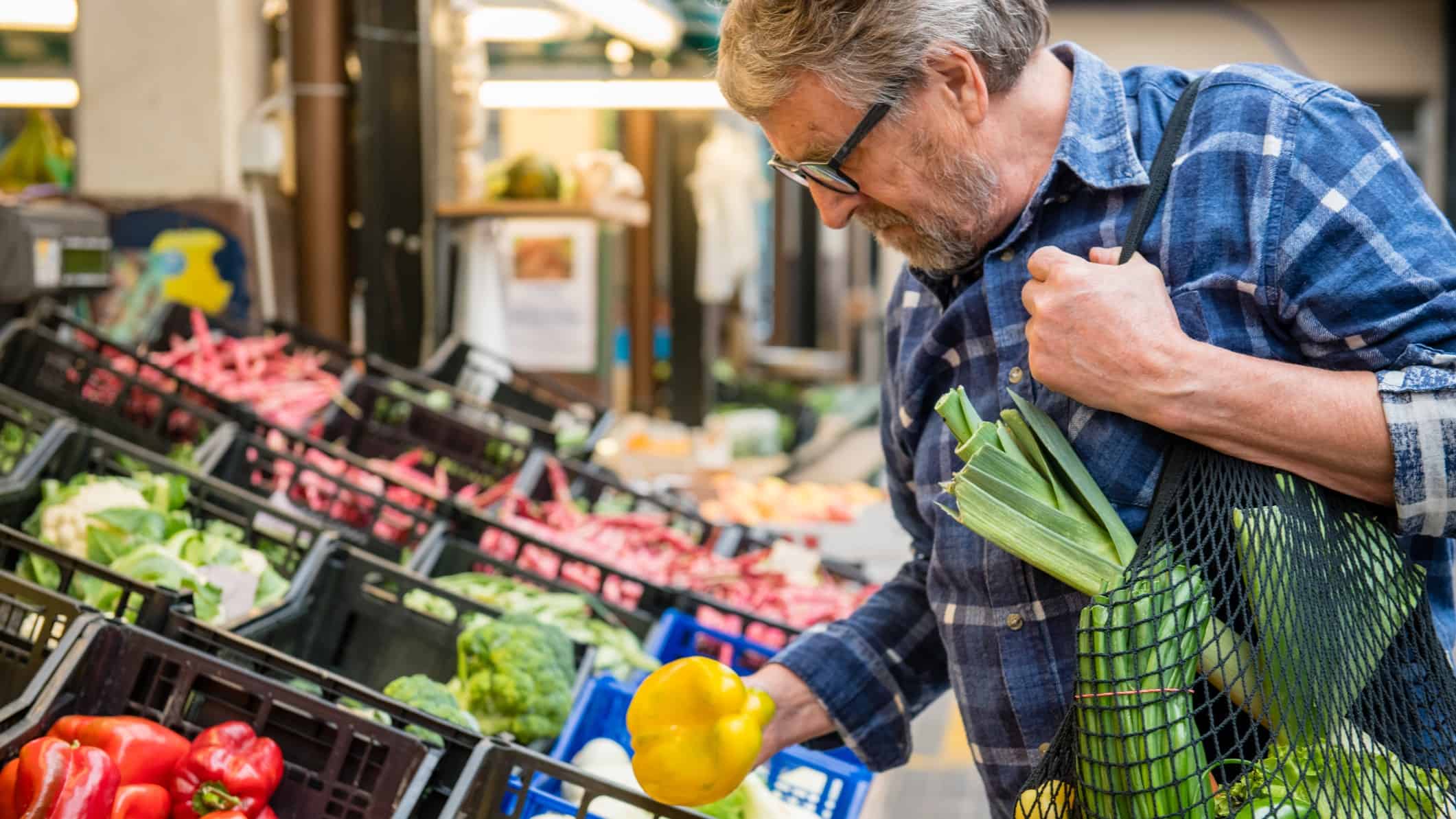 a man inspects a capsicum while holding an eco-friendly green string bag in a supermarket produce aisle.