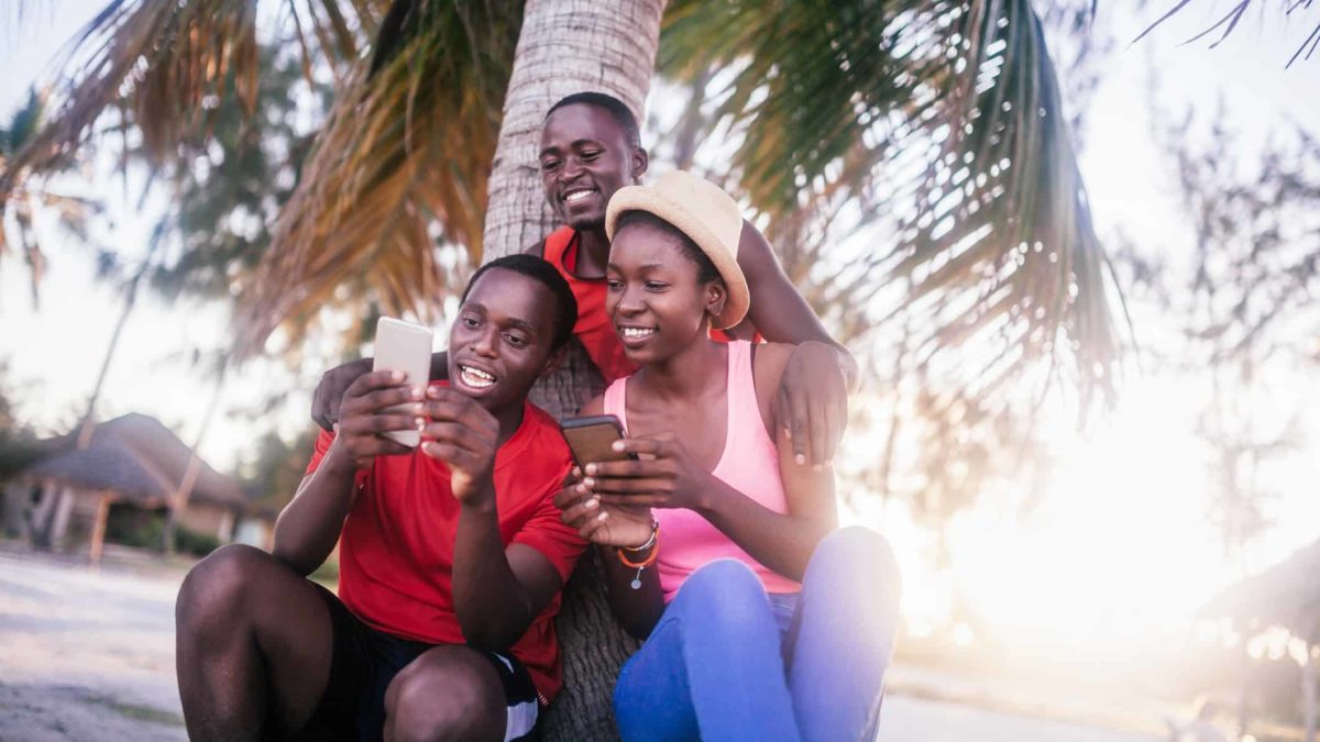 a group of islander residents look at their mobile phones under a palm tree.