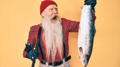 a fisherman with a long beard makes a crazy widemouthed face at a large salmon held by the tail in one of his hands.