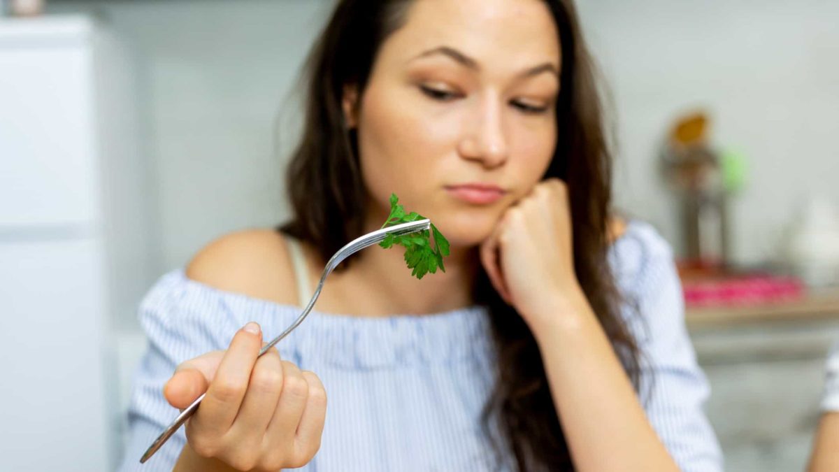 a sad woman holds a green vegetable on her fork and looks unhappy while propping up her chin with her hand.