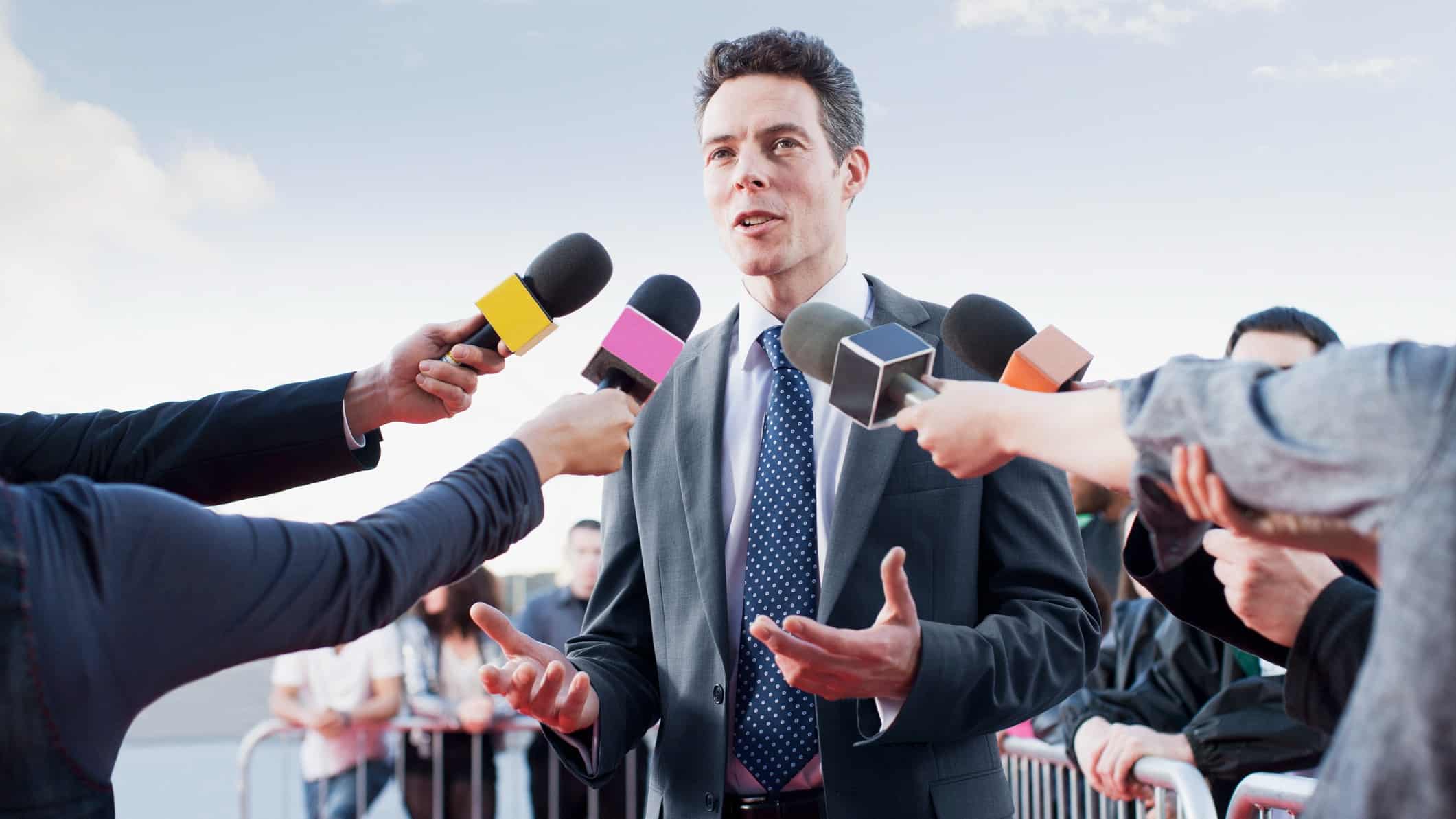 A man giving an interview before several handheld media microphones.