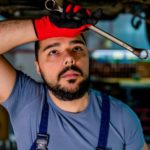 A mechanic wipes his forehead under a car with a tool in his hand and looking at car parts.