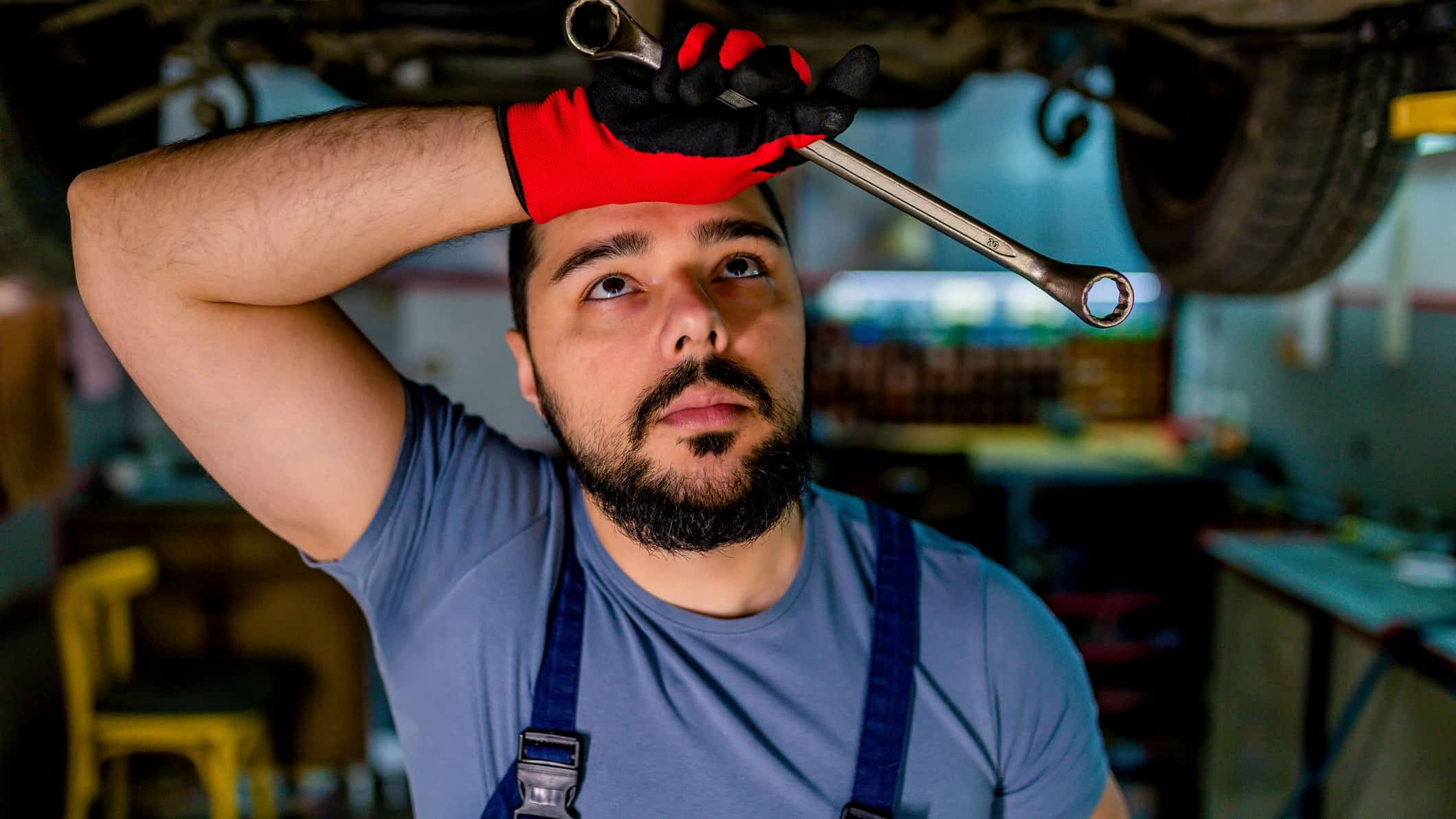 A mechanic wipes his forehead under a car with a tool in his hand and looking at car parts.
