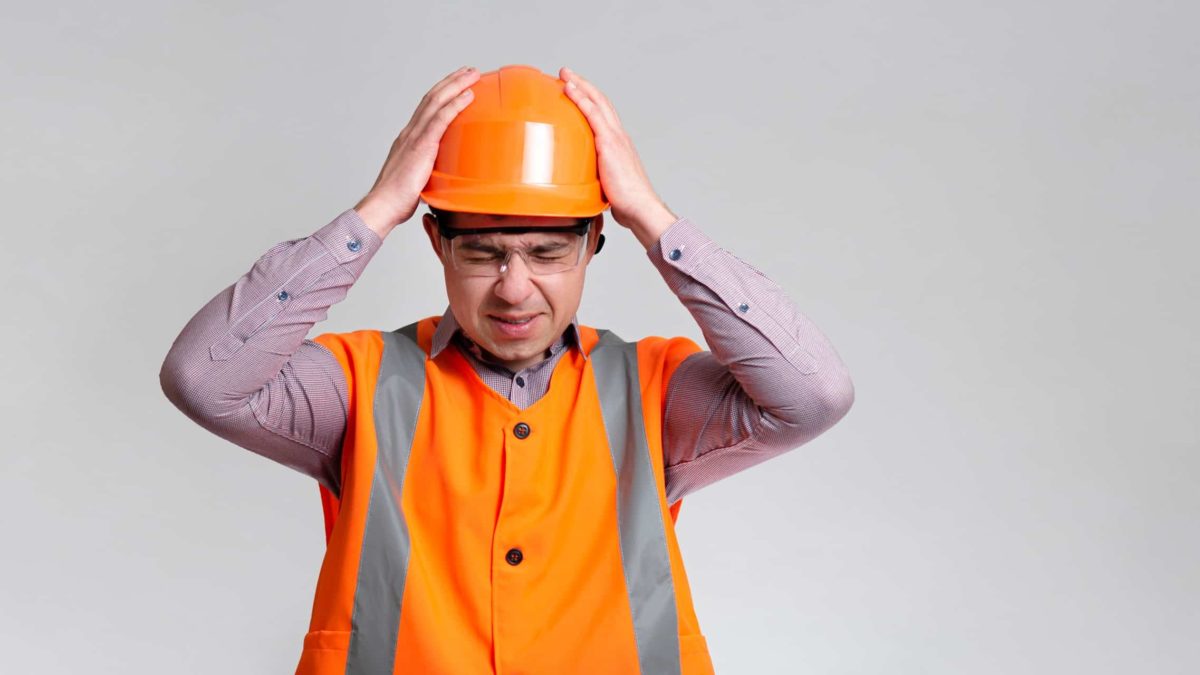 a builder wearing a hard hat and a safety high visibility vest closes his eyes and puts his hands on his head as if receiving bad news.