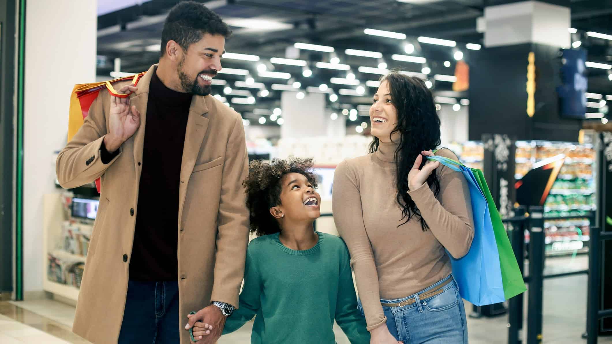 a family with shopping bags walks inside a shopping mall with shops in the background.