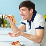 a happy man eats pizza in his kitchen with a long string of cheese between the pizza slice in his hand and in his mouth.