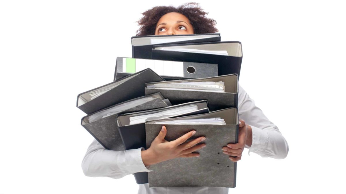 a woman struggles to hold a large pile of folders and documents with only her eyes appearing over the top of the pile.