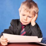 sad child holds paper and leans with head in hand near a computer looking downcast.