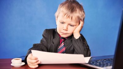 sad child holds paper and leans with head in hand near a computer looking downcast.