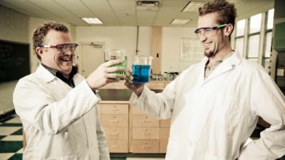 two chemists celebrate by jokingly clinking two containers of chemicals while they wear white laboratory coats and protective glasses in their lab.