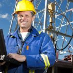 Happy miner using a computer at a mine, oil, or gas site with rigging in the background.