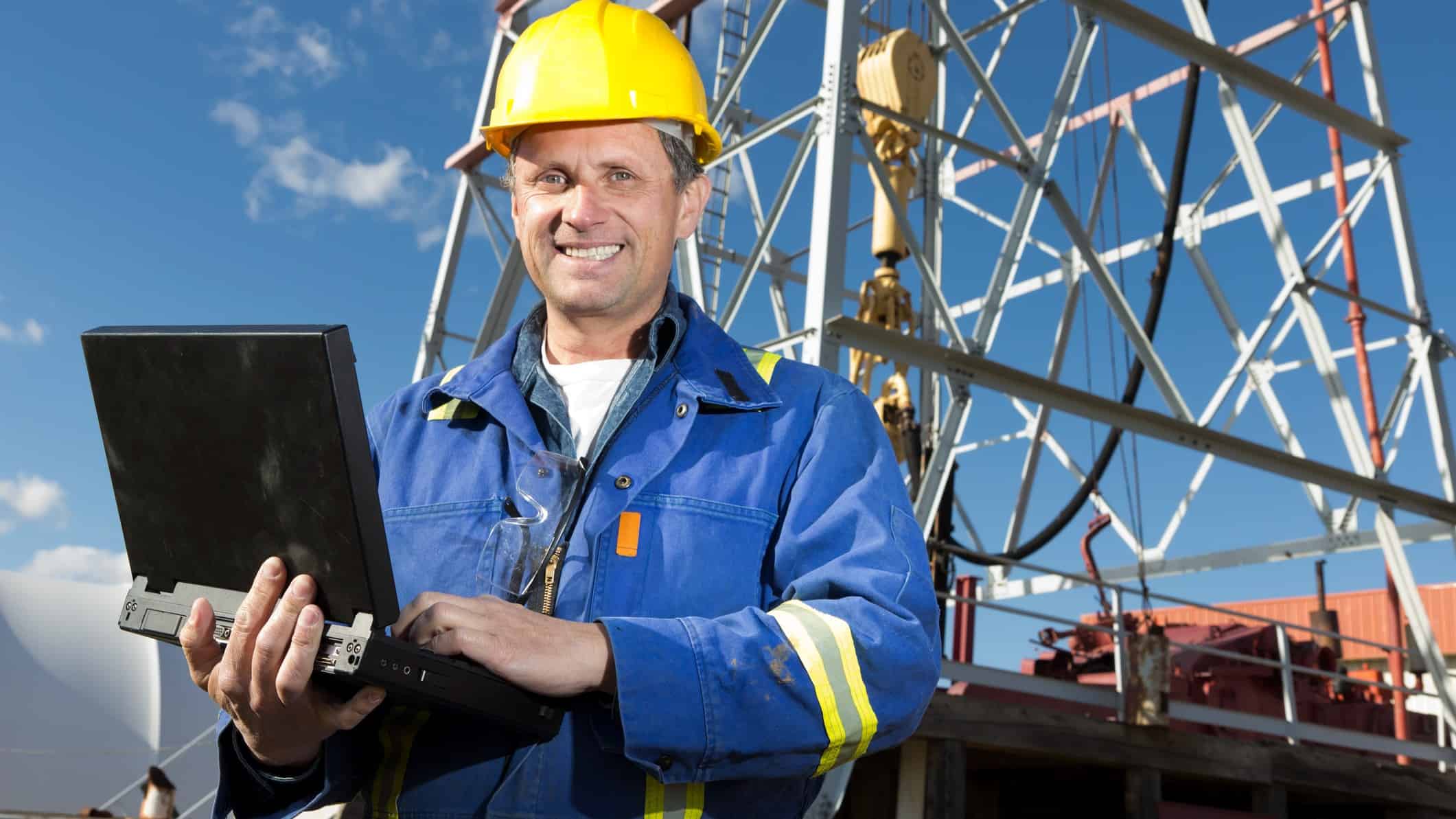 happy miner using a computer at a mine, oil or gas site with rigging in the background.
