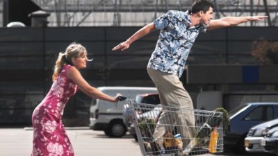 a woman pushes a man standing in a shopping trolley pointing ahead far off into the distance.