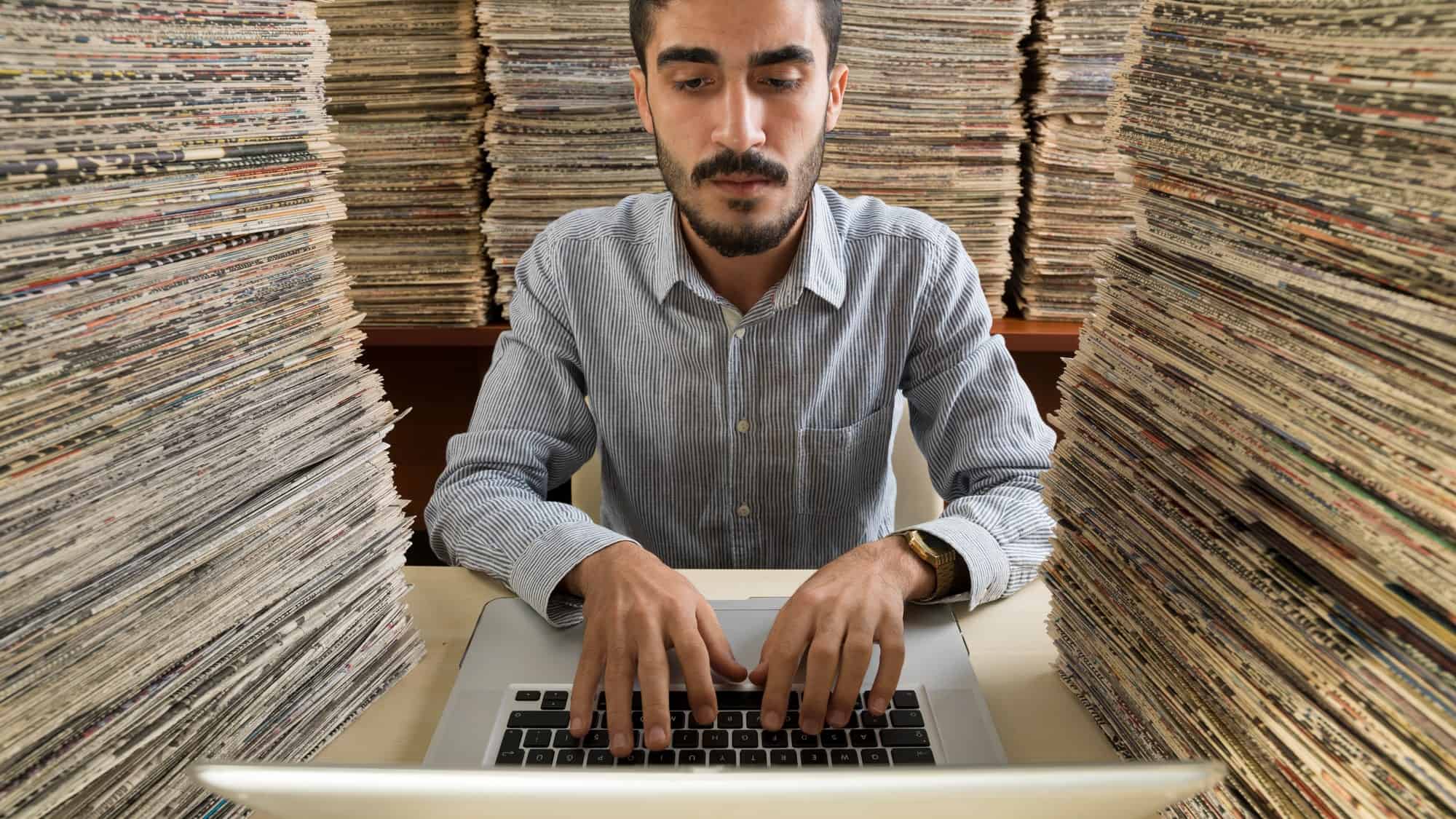 a man sits at a computer amid piles of papers to each side and behind him