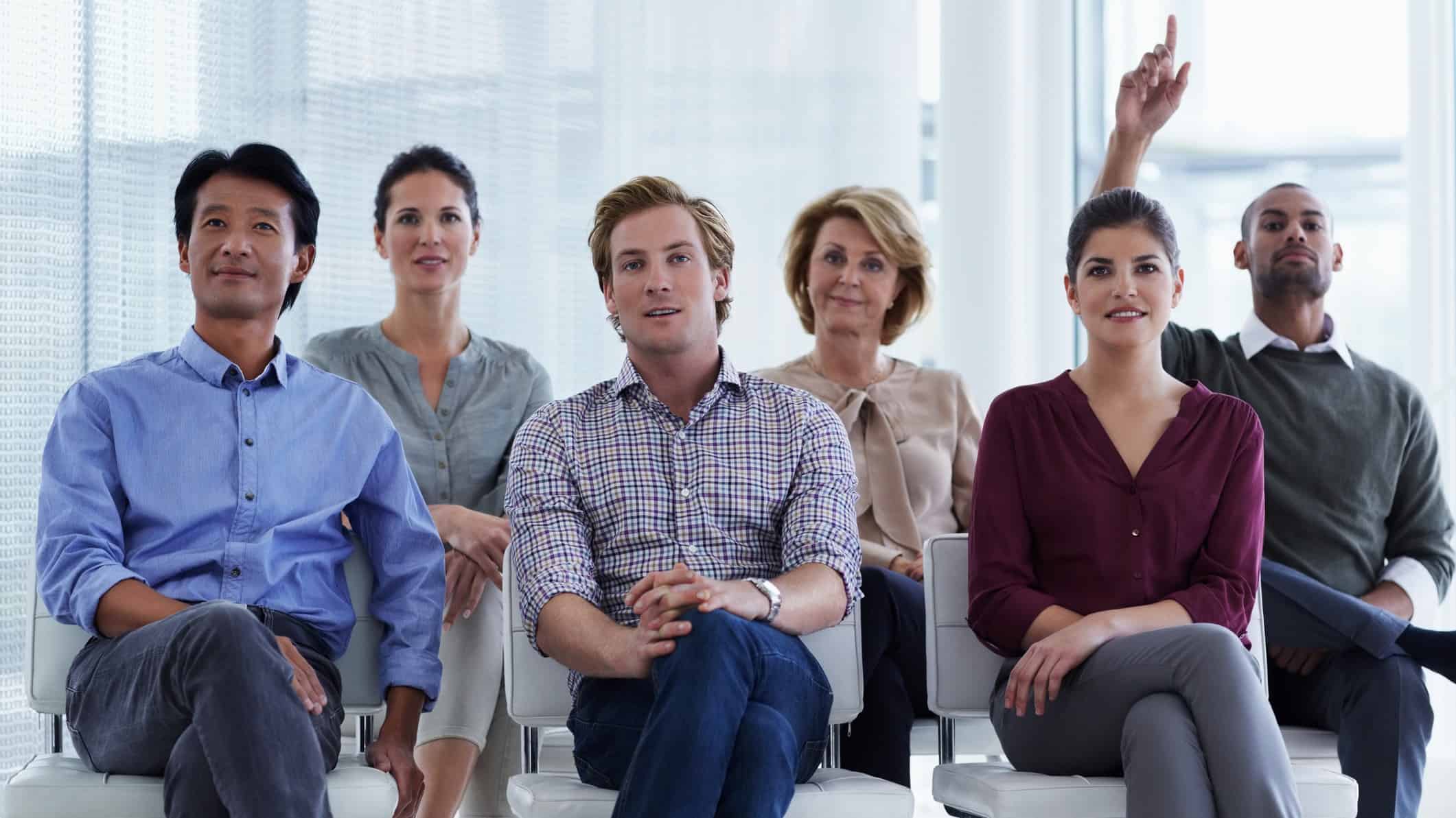 People sitting in rows in a meeting with one person holding their hand up as if to ask a question.