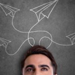 A man stands before a chalk board with line drawings of paper planes with various curling flight trajectories and paths.