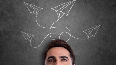 A man stands before a chalk board with line drawings of paper planes with various curling flight trajectories and paths.