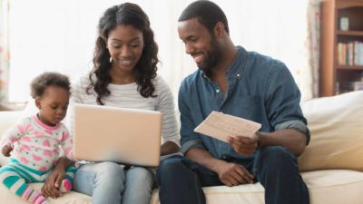 a couple and their baby sit together at their computer carrying out digital transactions and smiling happily.