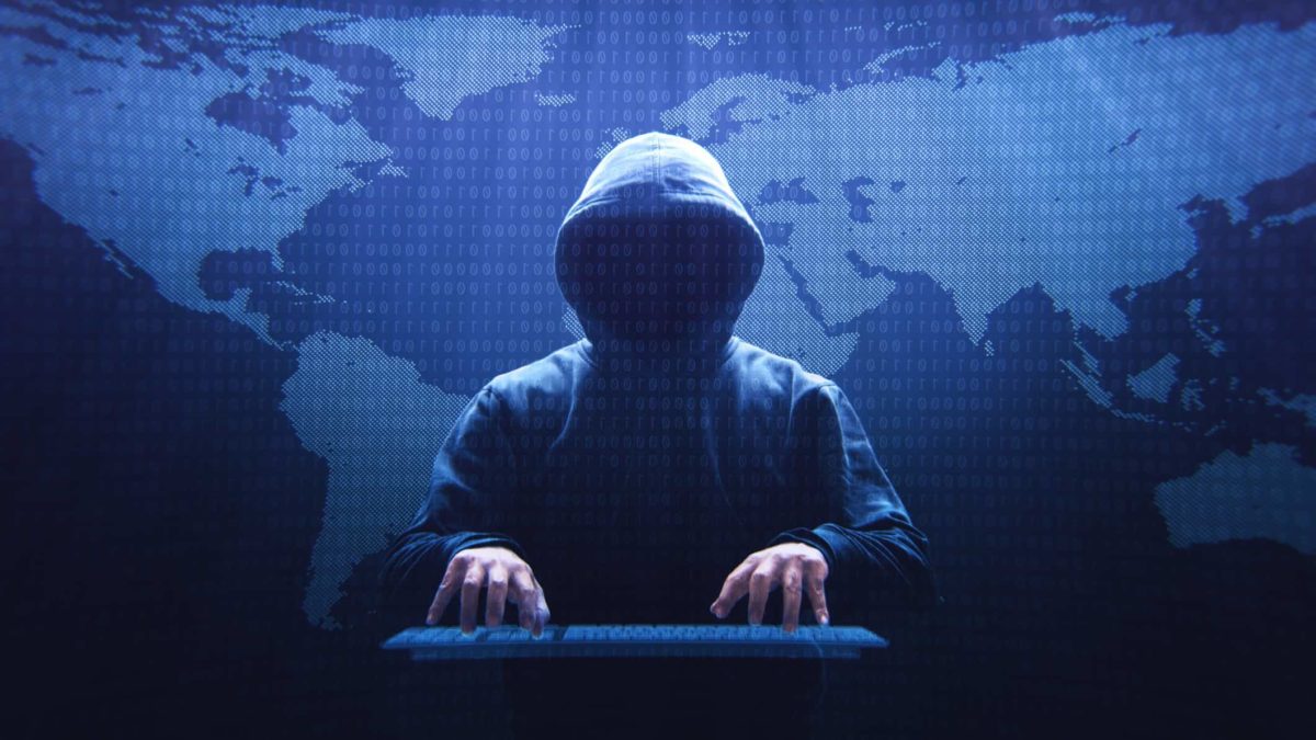 A hooded person sits at a computer in front of a large map of the world, implying the person is involved in cyber hacking.