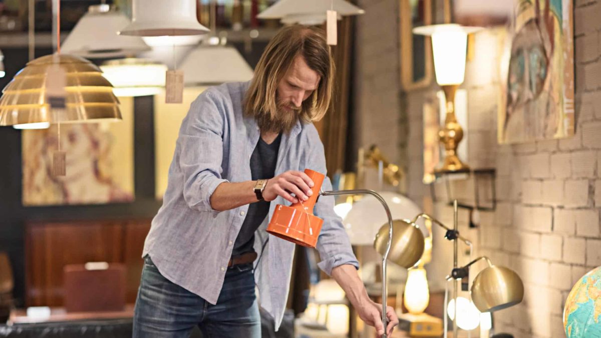 a man adjusts a desk lamp in a lighting store with an assortment of lamps and ceiling lights surrounding him.