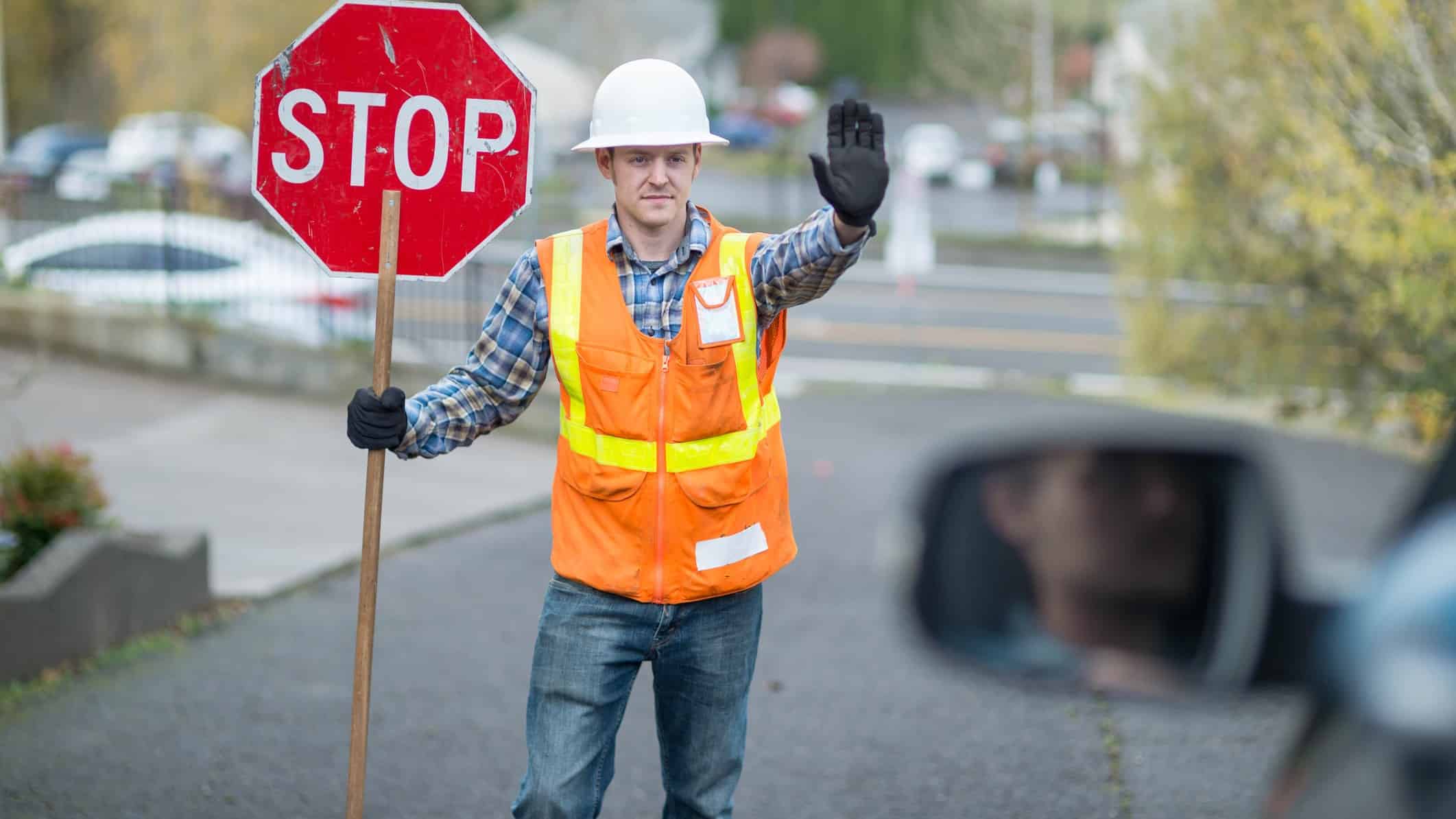 a man in a hard hat, high visibility vest and gloves holds a stop sign and holds up a hand in a halt gesture on a road.