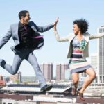 two people celebrating good news high five each other while jumping in the air with a city landscape in the background.