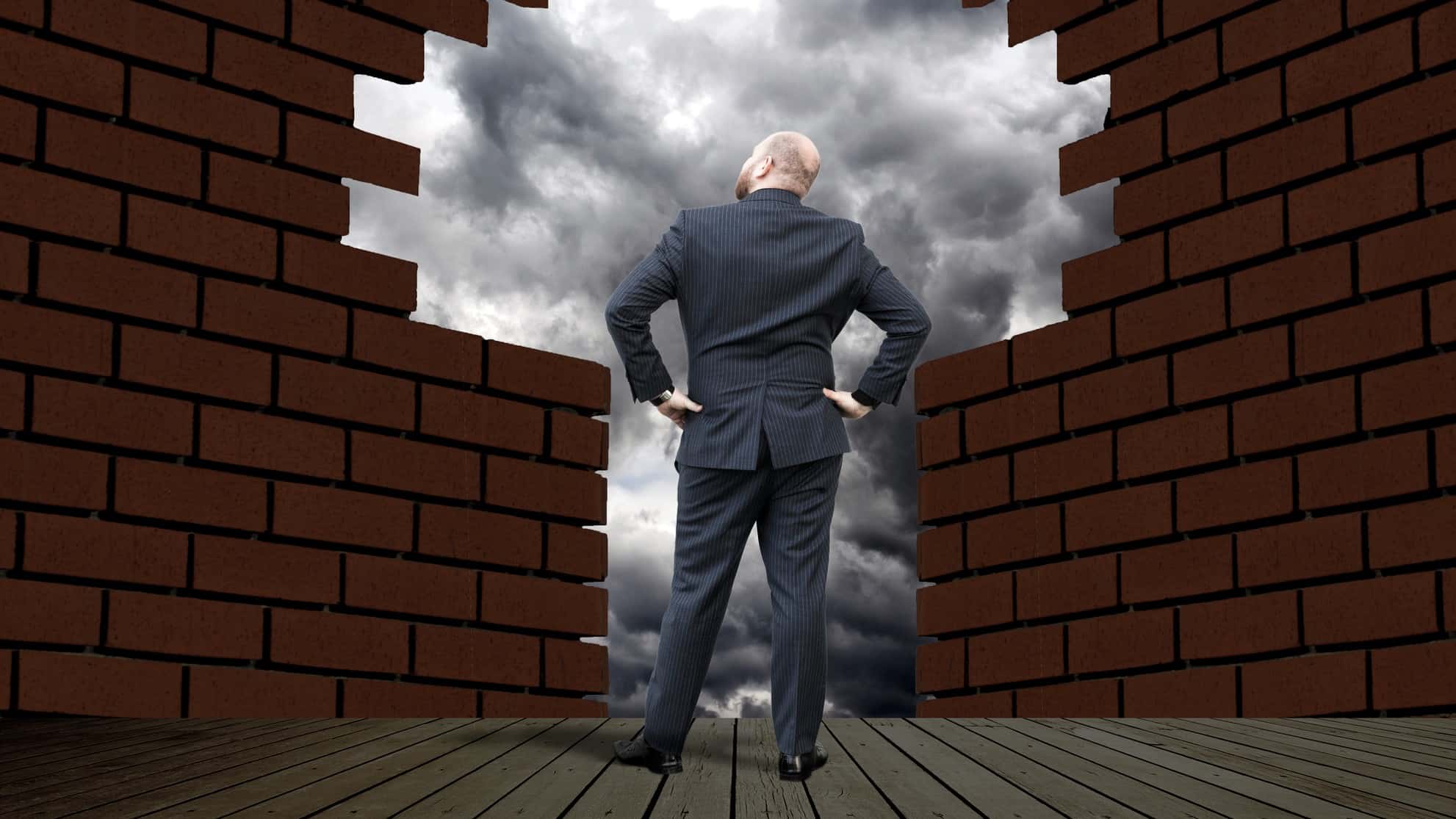 a man peers through a broken brick wall to see grey clouds gathering beyond it