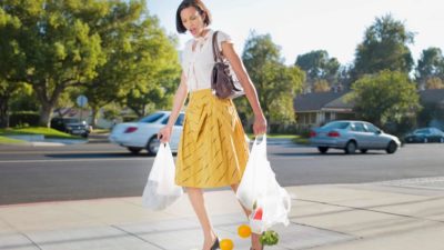 a woman walking along the street with plastic shopping bag makes an annoyed, angry expression as fruit and vegetables drop from a split in one of her bags.