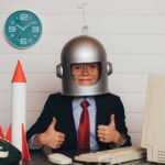 Child wearing a space helmet and sitting with thumbs up next to two toy rockets on a desk with a computer, keyboard and mouse.