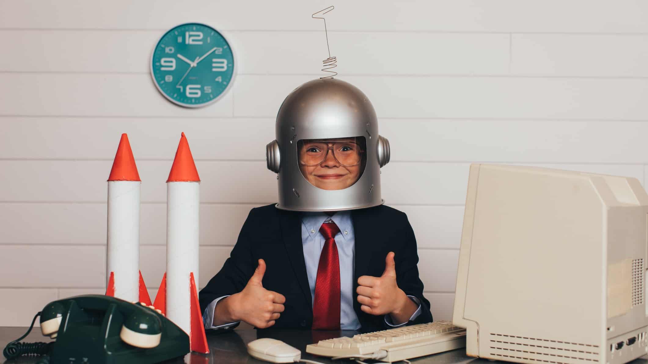 Child wearing a space helmet and sitting with thumbs up next to two toy rockets on a desk with a computer, keyboard and mouse.